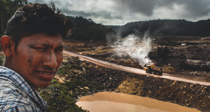 A member of the Mundukuru tribe looks into the camera, in the background construction of a dam is taking place.