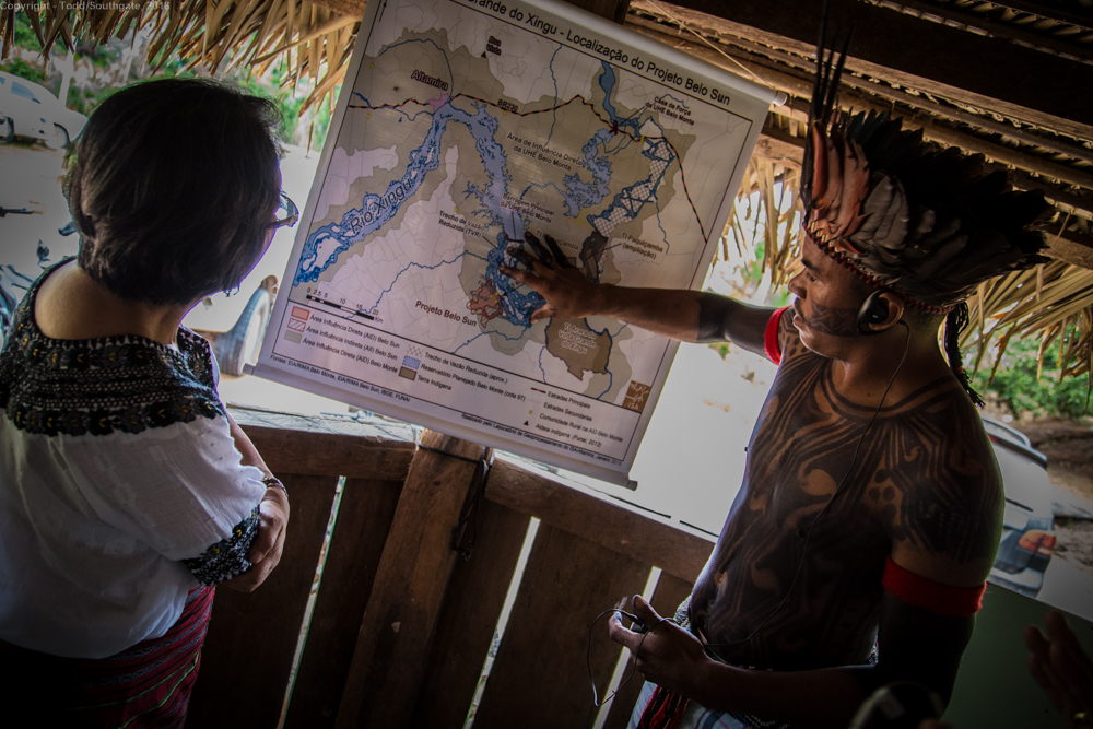 An Indigenous warrior in traditional dress points to a map of a river, while a woman to his left watches. 