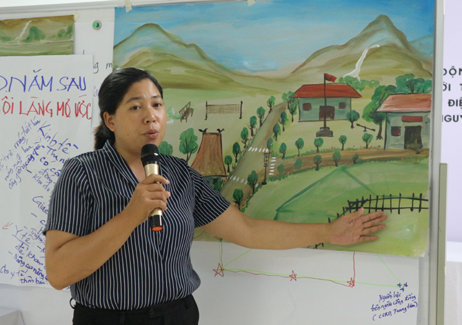 A woman holding a microphone gestures towards a poster of a town. 