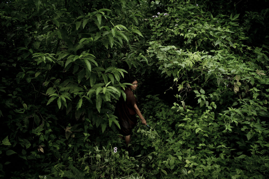 A woman walking into a dense area of greenery, looking back at the camera. 
