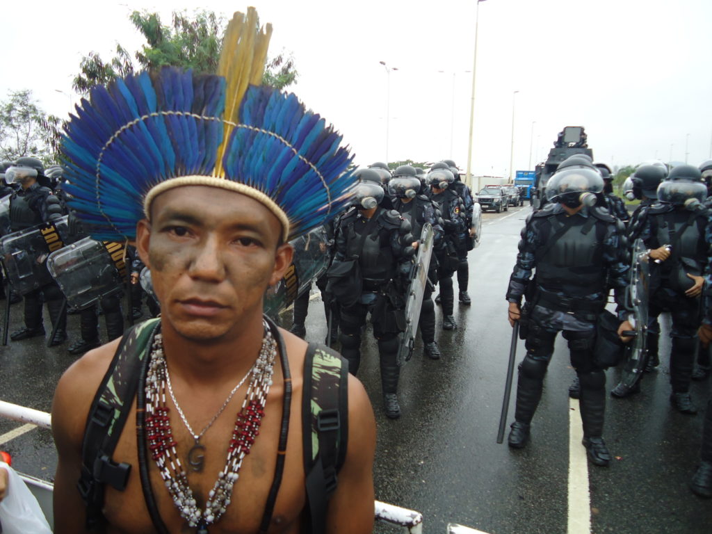 An indigenous leader stares into the camera with rows of police in riot gear standing behind him. 
