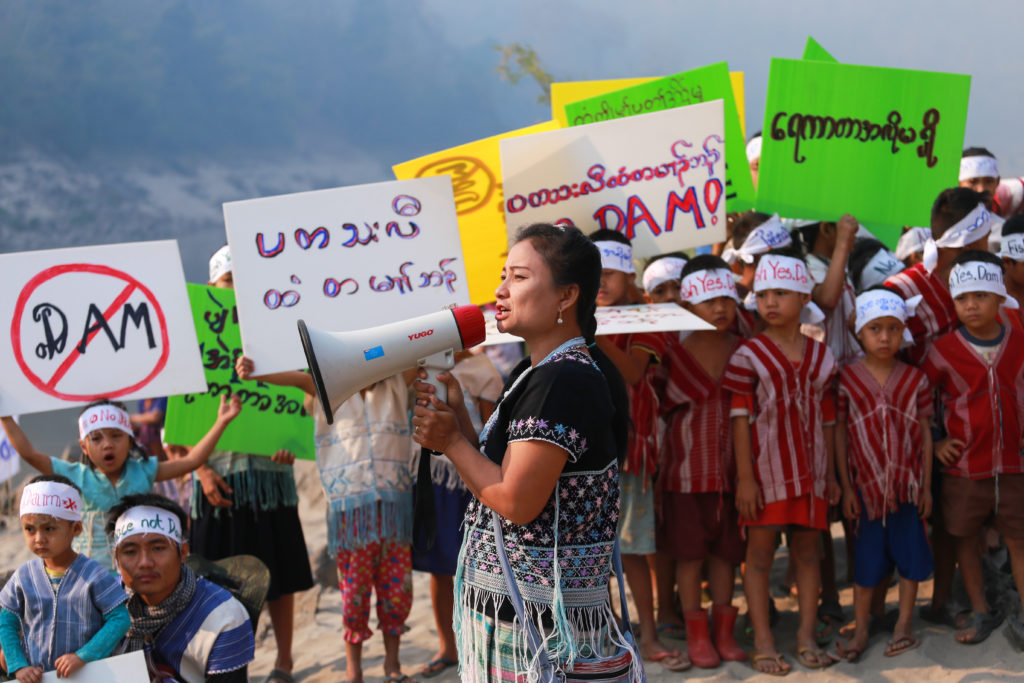 A woman speaks into a megaphone at a protest. There are children in the background holding posters. 