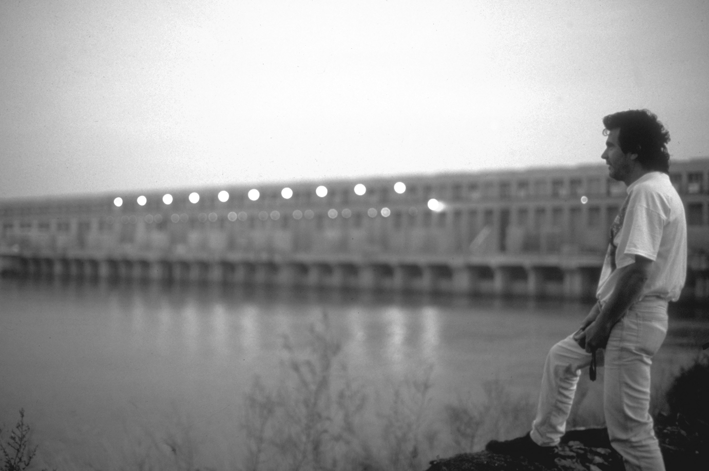 Black and white photo, Elías Díaz Peña Echegure over looks a river as the lights of a city twinkle in the background.