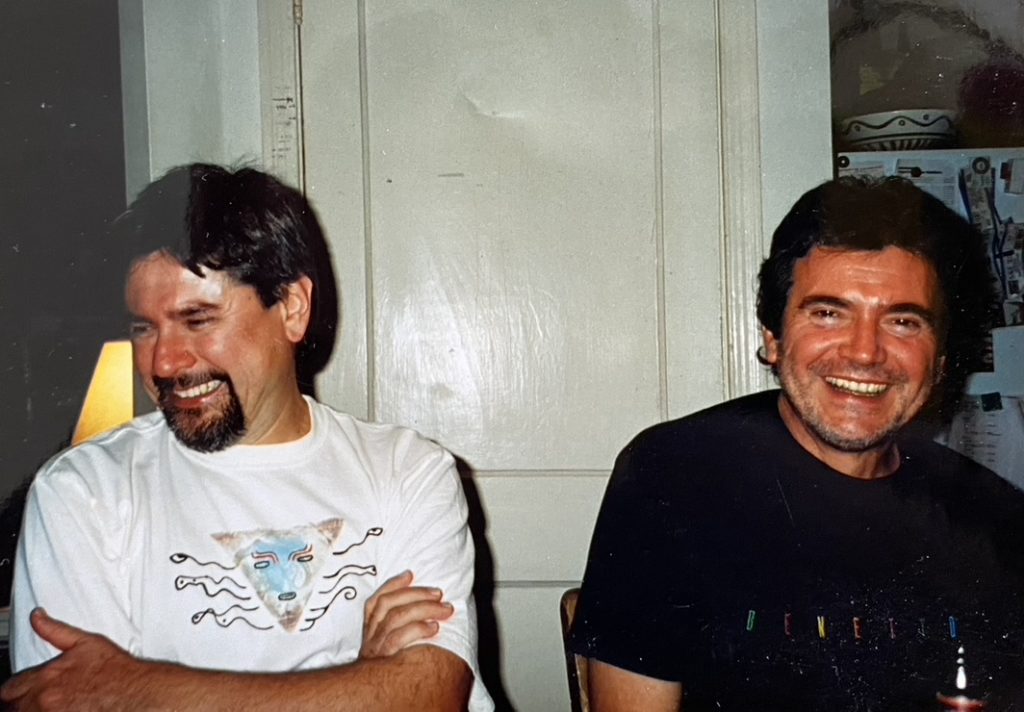Elías, wearing a black shirt, is sitting and smiling into the camera. To his right, partner Oscar Rivas sits, wearing a white shirt, crossing his arms, and smiling off to the side. 