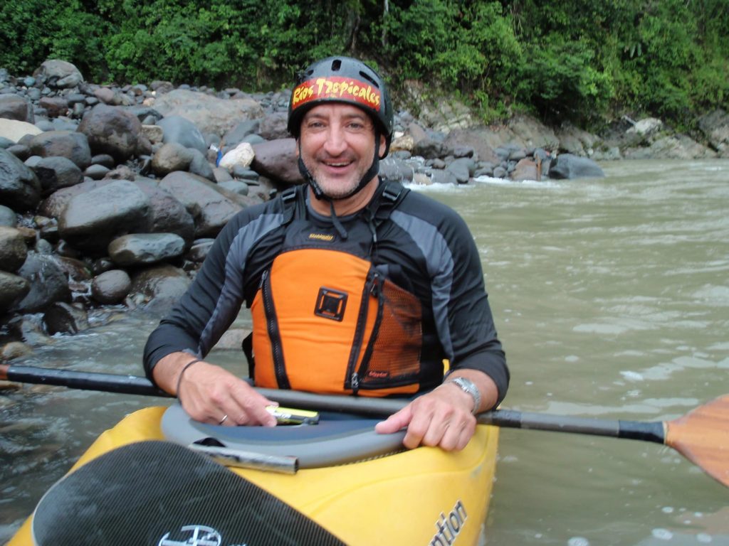 Rafa kayaking with his whitewater adventure company, Rios Tropicales. He smiles at the camera. The shore behind him is lined with green foliage and rocks.